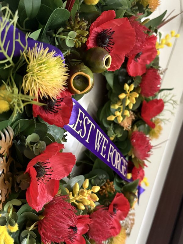 A zoomed in photo of a Remembrance Day wreath featuring yellow natives flowers, Poppies and a purple sash with the words ‘Lest We Forget’ in gold text.