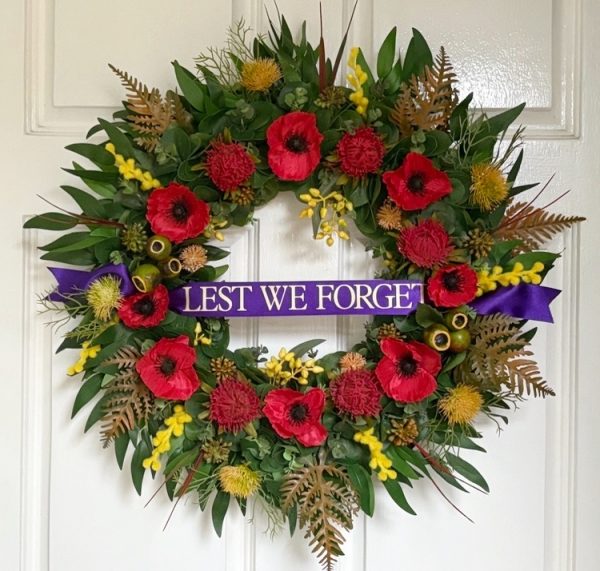 A Remembrance Day wreath on a white door decorated with Poppies, Waratahs, Wattle and Native Flowers with Eucalyptus and Autumn leaves around the circumference of the wreath. It has a purple sash across the front with the words ‘Lest We Forget’ in Gold Text.