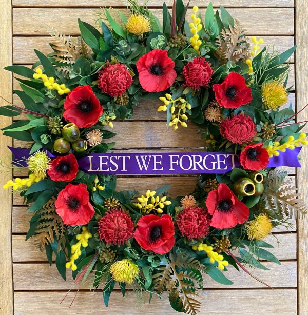 Remembrance Day handcrafted wreath featuring poppies, Waratahs, Wattle, Native yellow flowers and Gum nuts. The Eucalyptus and autumn leaves spread out around the circumference. It features a purple sash across the front with the words ‘Lest we forget’ in gold text.