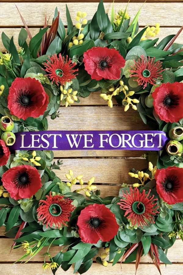 An ANZAC wreath featuring red poppies, red native flowers, gum nuts and eucalyptus leaves with long red spiky leaves, gum leaves, yellow flowers and wattle around the perimeter of the wreath.