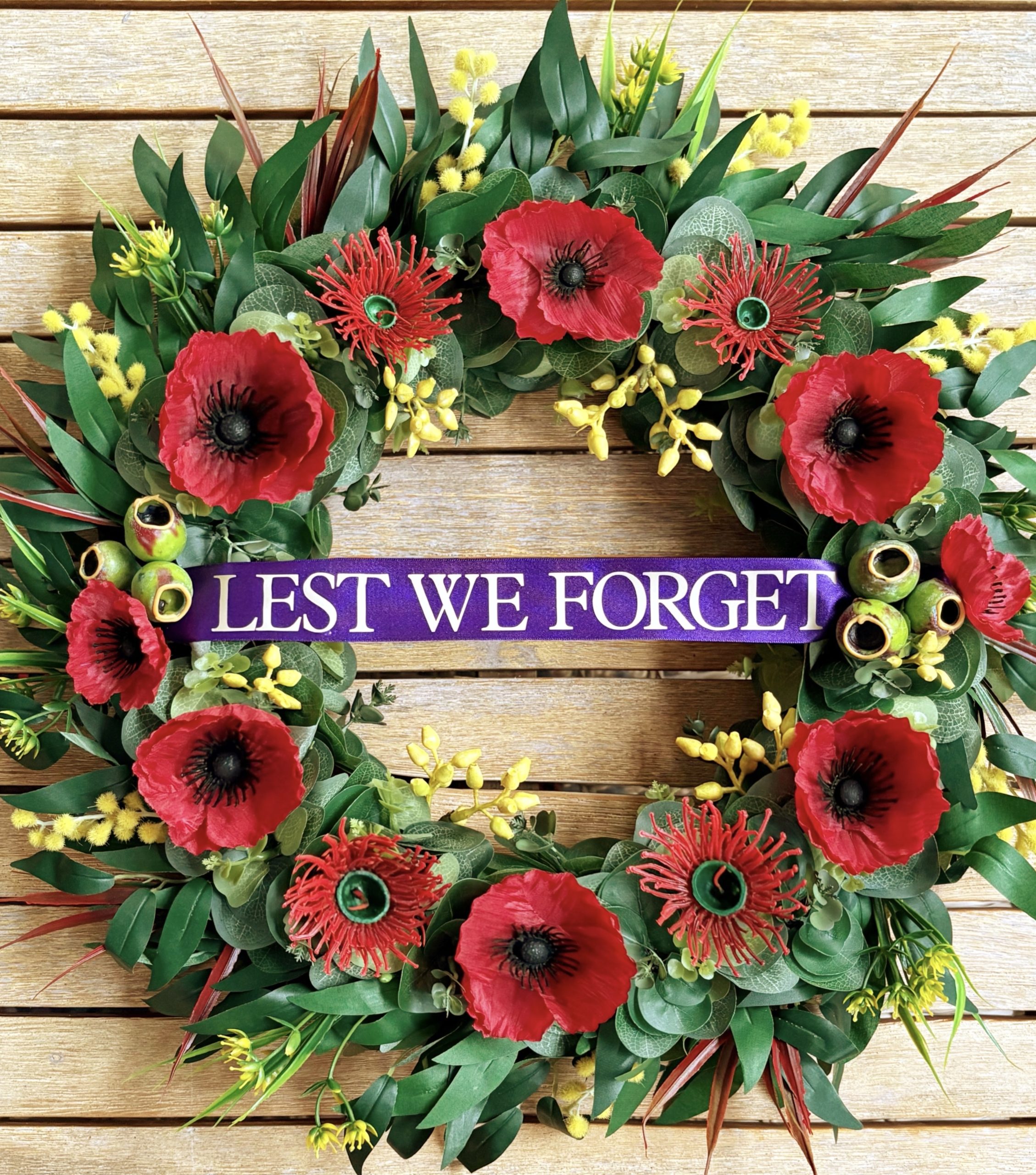 ptg_temp_save An ANZAC wreath featuring red poppies, red native flowers, gum nuts and eucalyptus leaves with long red spiky leaves, gum leaves, yellow flowers and wattle around the perimeter of the wreath.