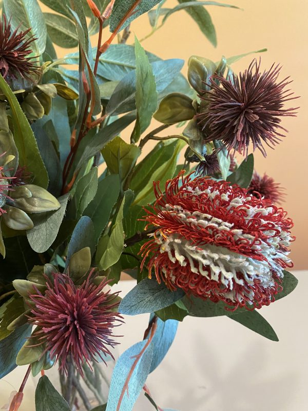 Close up of a red banksia flower, red thistle flowers with eucalyptus leaves behind it.