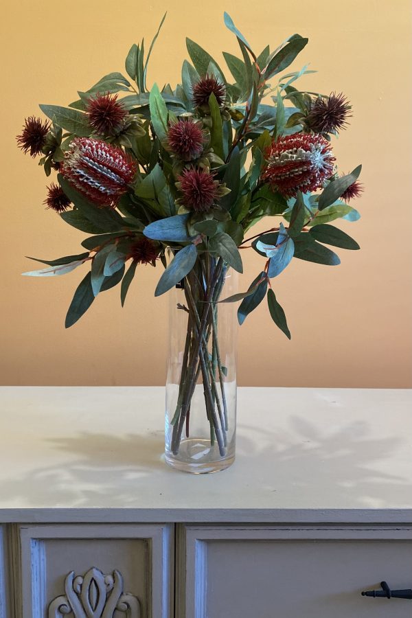 A large table arrangement of banksias, red thistle flowers and bushels of eucalyptus leaves in a clear vase on a white dresser